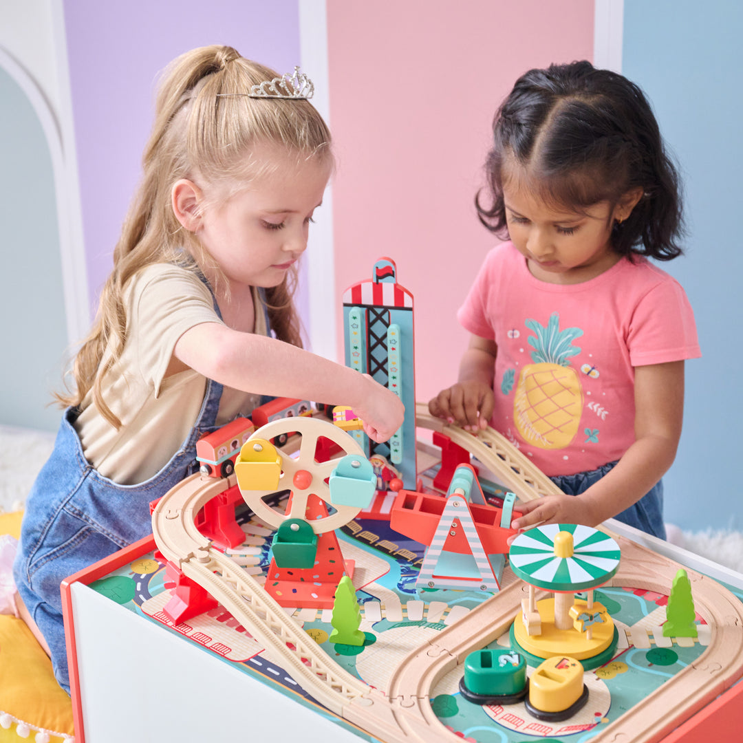 Two children playing together at a wooden carnival-themed train table with colorful tracks and rides.