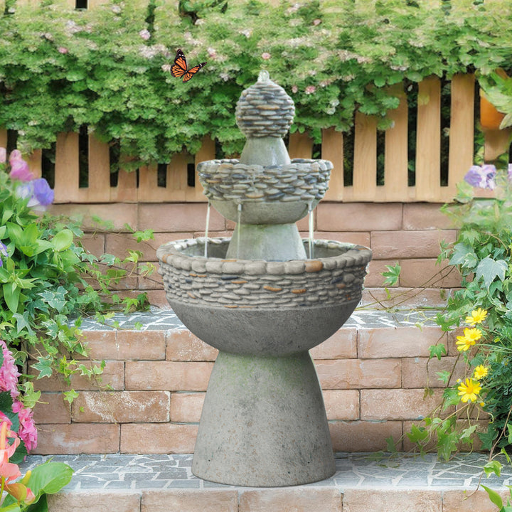A 3-tiered birdbath style water fountain with pebble accents in front of a brick and wood wall