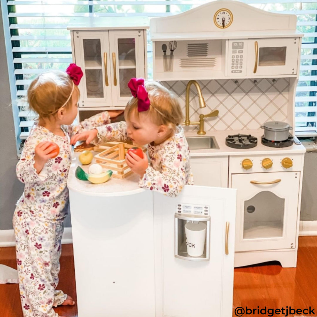 Two children in floral pajamas play with a toy kitchen set, featuring a stove, sink, and accessories. Both have pink bows in their hair.