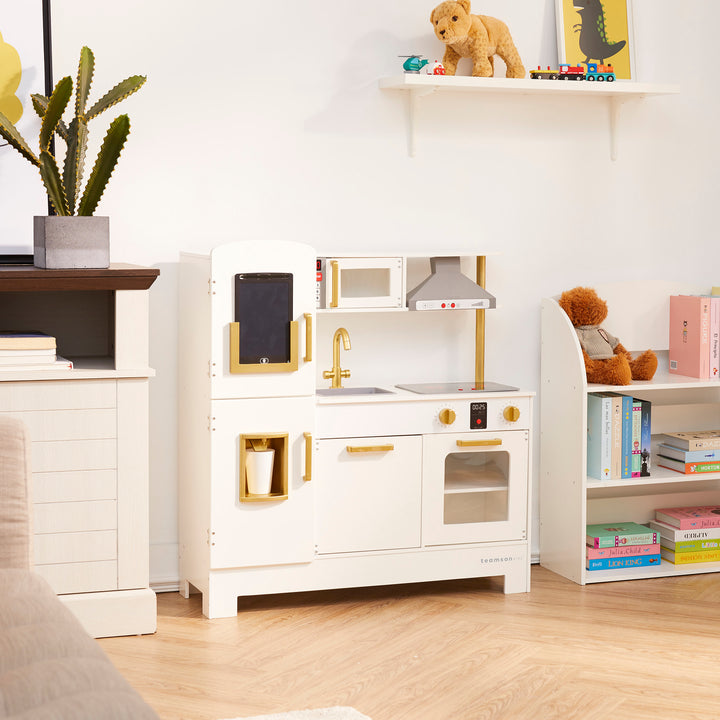 Teamson Kids All-in-One Little Chef Munich Play Kitchen with Lights and Sound Effects, White against a white wall. The kitchen is next to a bookshelf that has a teddy bear on top.